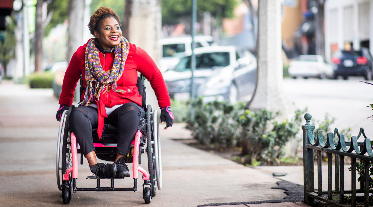 A young black disabled woman with a wheelchair and a bright colored sweater and her Asian friend walk around the city.