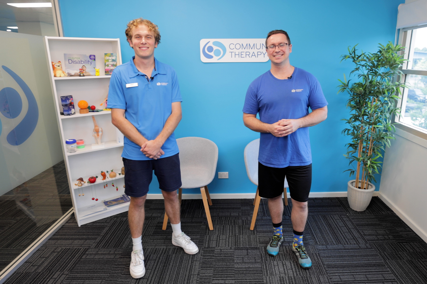 Scott and Harry standing and smiling in a bright exercise room, ready to lead a Senior Strong session. A bookshelf with therapy items and a Community Therapy sign are in the background.